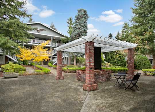 A landscaped outdoor gathering area here at Colonial Square featuring a brick pergola with a built-in grill, a small table with chairs beneath the covered structure, and a paved courtyard surrounded by mature evergreen trees, colorful seasonal foliage, manicured shrubs, and two-story apartment buildings with exterior walkways.
