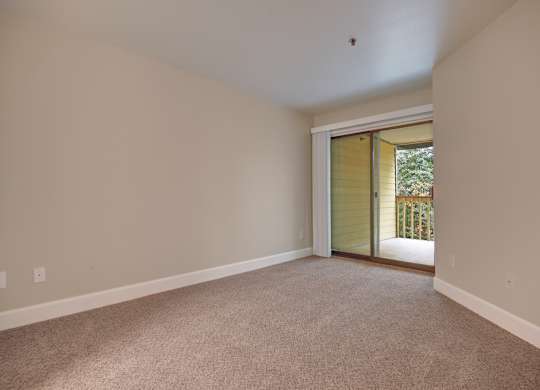 A spacious bedroom here at Forest Greens featuring soft carpet flooring, neutral wall tones, and an open layout that offers flexible furniture placement. The room includes a sliding glass door with vertical blinds that opens to a private balcony with a railing and leafy outdoor views, allowing natural light to brighten the space and create an airy, comfortable atmosphere.