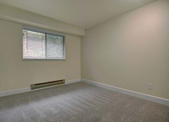 A bright bedroom here at Forest Greens featuring plush carpet flooring, soft neutral wall tones, and a simple open layout that allows for flexible furniture placement. The space includes a window with white blinds that brings in natural light, a baseboard heater below the window, clean white trim, and an uncluttered design that creates a calm, comfortable atmosphere.