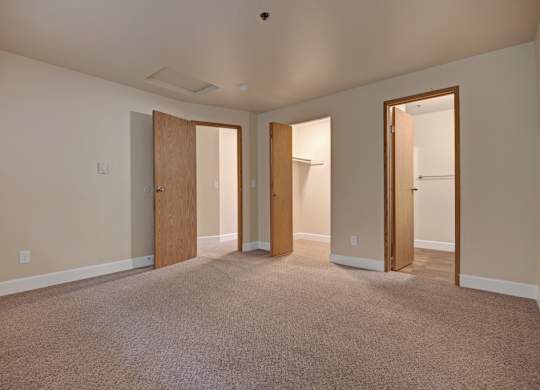 A spacious bedroom here at Forest Greens featuring plush carpet flooring, neutral wall tones, and a wide open layout that allows for flexible furniture placement. The room includes multiple doorways leading to a walk-in closet and an attached bathroom, clean white trim, simple overhead lighting, and a calm, uncluttered design ideal for comfortable everyday living.