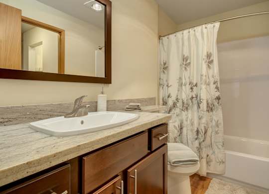 A clean bathroom here at Forest Greens featuring a wood vanity with a light stone-style countertop and integrated sink beneath a framed mirror. A bathtub-shower combination with white wall surround and patterned shower curtain sits beside the vanity, while wood-style flooring, folded towels, and neutral finishes create a tidy, welcoming space.