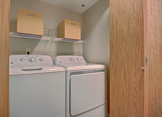 A convenient in-home laundry closet here at Forest Greens featuring a side-by-side washer and dryer tucked behind wood-paneled doors. White wire shelving above provides storage space for supplies, while neutral walls and a compact layout keep the area functional and easy to access.