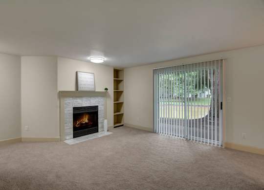 A spacious living room here at Island Park featuring soft neutral carpeting, a tiled fireplace with a simple mantel, and built-in open shelving for storage or décor. Vertical blinds cover a sliding glass door that opens to a patio or outdoor view, allowing natural light to fill the room and highlight the open, flexible layout.