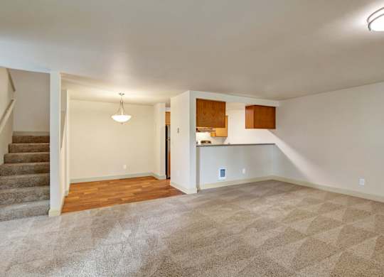 A spacious open living area here at Island Park featuring plush neutral carpeting, an open staircase with carpeted steps, and a dining nook with wood-style flooring and a hanging light fixture. The layout flows into a kitchen pass-through with upper cabinetry, creating an open, flexible space suitable for both living and dining.