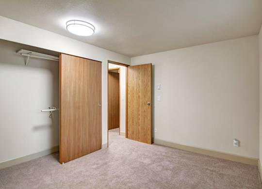 A simple unfurnished bedroom here at Island Park featuring soft neutral carpeting, an open closet with wood sliding doors and hanging shelves, and a doorway leading to the hallway. The room includes a large window with horizontal blinds allowing natural light to fill the space, offering a clean, flexible layout suitable for a bedroom or home office.