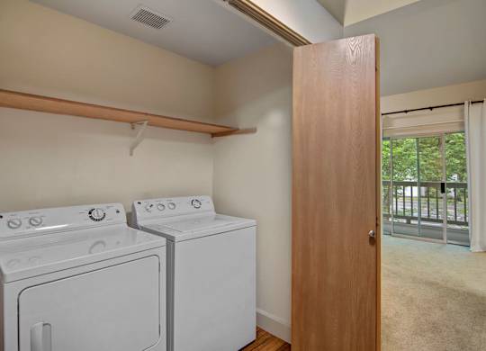 A convenient in-home laundry area here at Island Park featuring a full-size washer and dryer set beneath an open storage shelf, with wood-style flooring and neutral walls. The space opens to the living area through a wood door and sits near a sliding glass door that brings in natural light, creating a functional and easy-to-access layout.