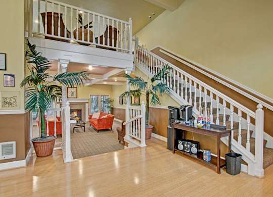 A spacious community lobby here at Island Park featuring a two-story layout with white railings, an open staircase, and warm wood flooring. Potted palm plants frame a cozy seating area with accent chairs and a fireplace in the background, while a refreshment station adds convenience to this inviting shared space.