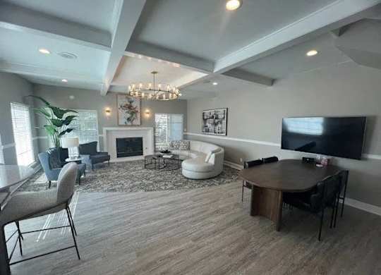A spacious clubhouse lounge here at Island Park featuring wide plank flooring, a coffered ceiling with recessed lighting, and a cozy seating area arranged around a fireplace. The room includes a curved sofa, accent chairs, wall art, a mounted television, and a round table with chairs, creating a welcoming space for relaxing or gathering.
