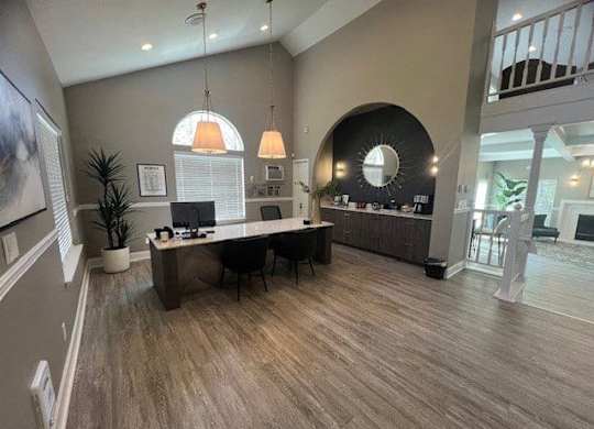 A welcoming leasing office here at Island Park featuring wood-style flooring, a central desk with computer stations and chairs, and modern pendant lighting overhead. The space includes a curved accent wall with a built-in counter, decorative mirror, greenery, and an open layout that connects to the clubhouse seating area beyond.