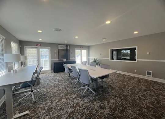 A modern meeting room here at Island Park featuring long conference tables with rolling chairs, patterned carpet flooring, recessed ceiling lighting, and neutral gray walls. The space includes large windows, glass double doors, a wall-mounted display, and a clean open layout designed for resident gatherings, meetings, or collaborative use.