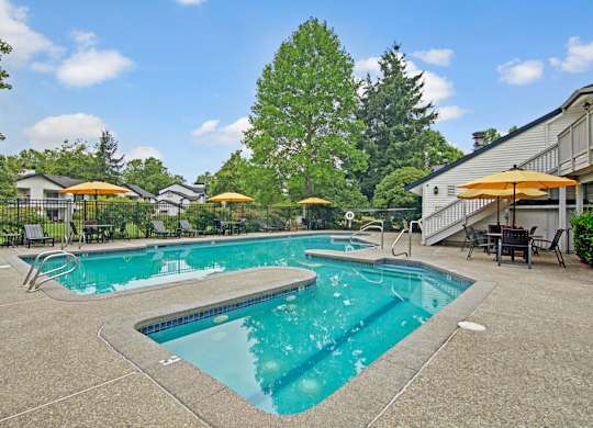 A refreshing outdoor swimming pool here at Island Park featuring a spacious concrete deck, a separate shallow pool area, and multiple lounge chairs with bright umbrellas. The fenced pool area is surrounded by mature trees, landscaped greenery, and nearby community buildings, creating a relaxing setting for residents.