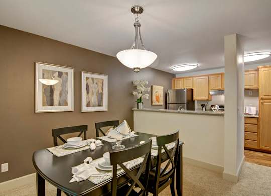 A welcoming dining area here at Island Park featuring a dark wood dining table set for four beneath a modern pendant light, with framed abstract artwork on an accent wall. The space flows into a kitchen with warm wood cabinetry, a breakfast bar, and coordinated lighting, creating a cohesive and inviting layout.