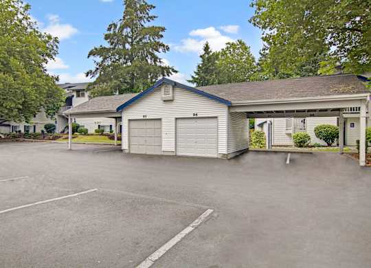 A covered parking and garage area here at Island Park featuring numbered private garages and open carport spaces with paved parking, surrounded by mature trees, landscaped greenery, and nearby apartment buildings under a bright sky with scattered clouds.