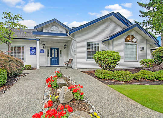 A welcoming leasing office exterior here at Island Park featuring a white clubhouse-style building with blue trim, a covered entry, and a central walkway lined with red flowers, decorative rocks, manicured shrubs, and green lawn under a bright blue sky with scattered clouds.