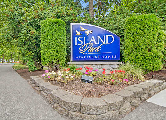 A landscaped monument sign here at Island Park featuring a blue Island Park Apartment Homes sign set on a stone base, framed by manicured evergreen shrubs, leafy trees, and colorful flower beds along a sidewalk in a well-maintained residential setting.