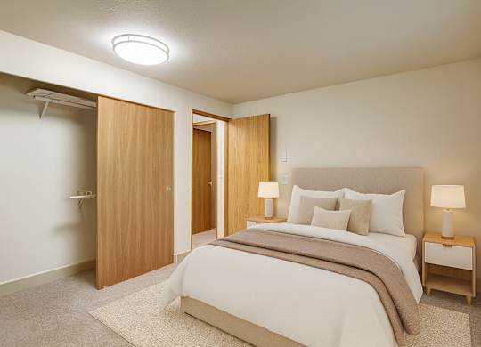 A calm bedroom here at Island Park featuring a bed with neutral bedding and layered pillows, a soft area rug over plush carpeting, and matching bedside tables with modern lamps. The space includes a sliding closet with wood doors, light-colored walls, and a wide window with blinds that brings in natural light.