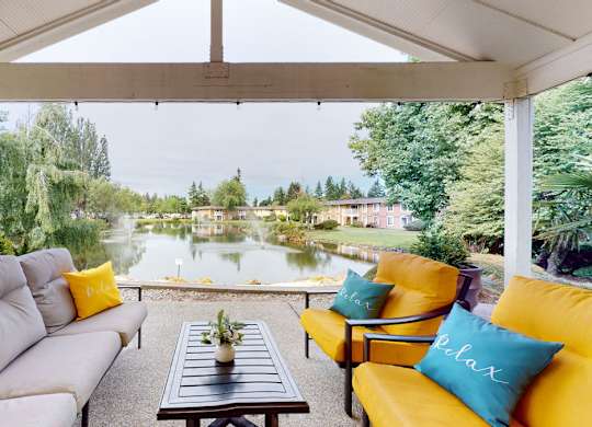 A covered outdoor seating area here at Lakeside featuring cushioned sofas and lounge chairs with colorful accent pillows arranged around a central coffee table, set beneath a peaked patio roof and overlooking a tranquil lake with fountains, mature trees, and surrounding apartment homes for a relaxing waterfront view.