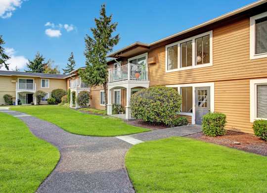 A landscaped courtyard here at Lakeside showcasing two-story apartment buildings with warm-toned siding, private balconies and patios, neatly trimmed shrubs, mature trees, and lush green lawns, with a winding paved walkway leading through the community beneath a bright blue sky.