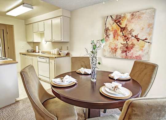 A cozy dining area here at Lakeside showcasing a round dining table set for four with upholstered chairs, neutral carpeting, and a decorative centerpiece, complemented by soft wall art and an adjacent kitchen with light cabinetry, ample counter space, and overhead lighting that creates a warm, inviting flow between spaces.