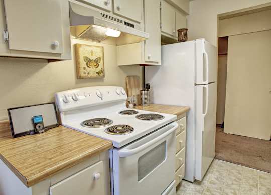 A cozy kitchen here at Lakeside featuring light-toned cabinetry, wood-style countertops, and a clean cooking area with a range, refrigerator, and overhead storage, accented by simple wall art, small countertop décor, and an open closet doorway that adds depth and functionality to the space.