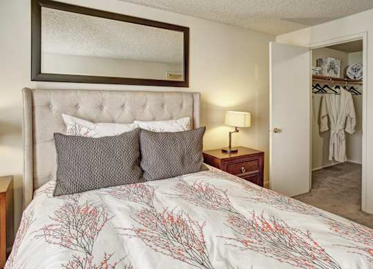 A cozy bedroom here at Lakeside featuring a tufted upholstered headboard with layered decorative pillows and patterned bedding, paired with a wooden nightstand and table lamp, a large framed mirror above the bed, plush carpet flooring, and an open closet with hanging space that adds functionality and storage.