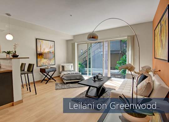 A stylish open living room here at Leilani on Greenwood featuring light wood-style flooring, a modern seating area with a sofa and lounge chair, and a sleek coffee table atop an area rug. The space opens to a contemporary kitchen with bar seating, while sliding glass doors bring in natural light and connect to an outdoor patio.