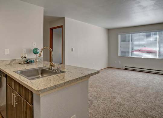 A spacious open living area here at Leilani on Greenwood featuring soft carpeted flooring, neutral walls, and a large window with blinds that brings in natural light. The space connects to a modern kitchen with a light stone countertop, undermount sink, and wood-style cabinetry, creating a comfortable and functional open-concept layout.