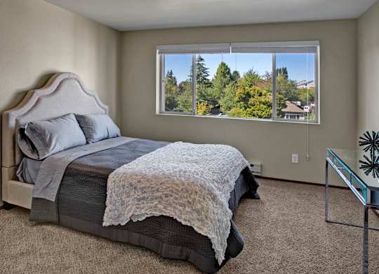 A bright bedroom here at Leilani on Greenwood featuring soft carpeted flooring, neutral walls, and a bed with layered gray and white bedding. A large window with blinds fills the room with natural light and offers leafy neighborhood views, while the open layout provides comfortable space for bedroom furnishings.