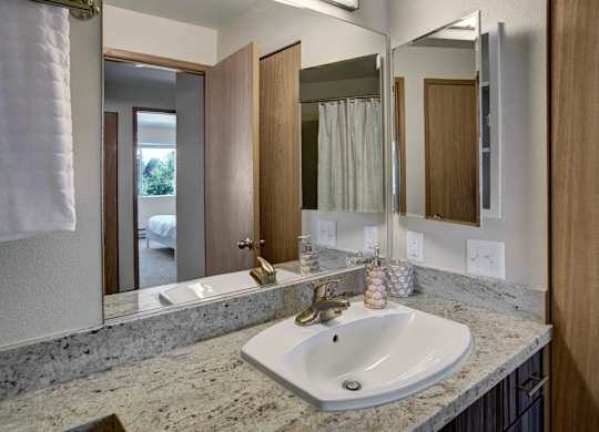 A clean bathroom here at Leilani on Greenwood featuring a light stone countertop with an integrated sink, a wood vanity with storage below, and a wide wall mirror with overhead lighting. Neutral walls, coordinating fixtures, and a nearby doorway create a bright, functional layout connected to the bedroom.