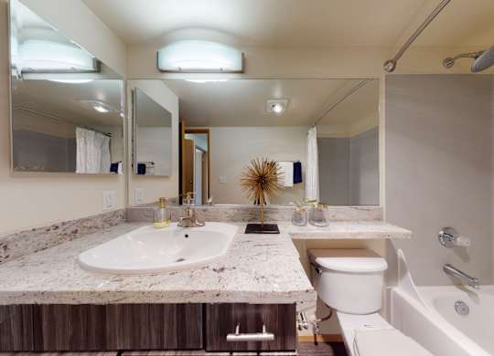 A well-appointed bathroom here at Leilani on Greenwood featuring a wood vanity with a light stone countertop, an integrated sink, and wide wall mirrors with overhead lighting. A toilet sits beside the vanity, while a bathtub-shower combo with a curtain completes the clean, functional layout.