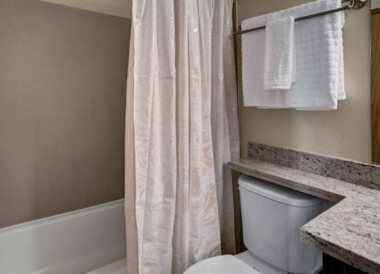 A simple bathroom here at Leilani on Greenwood featuring a bathtub-shower combo with a curtain, a toilet positioned beside a light stone countertop, and neutral wall finishes. A towel bar with folded towels and clean lines create a practical, easy-to-use space within the apartment.