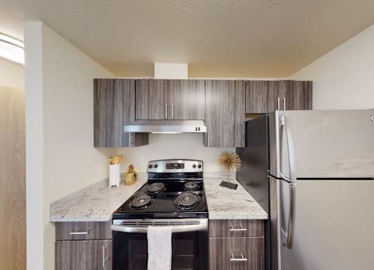 A modern kitchen here at Leilani on Greenwood featuring gray wood-style cabinetry, light stone countertops, and a full cooking area with a range, hood, and refrigerator. Upper and lower cabinets provide storage, while clean finishes, simple décor, and overhead lighting create a functional and contemporary kitchen layout.
