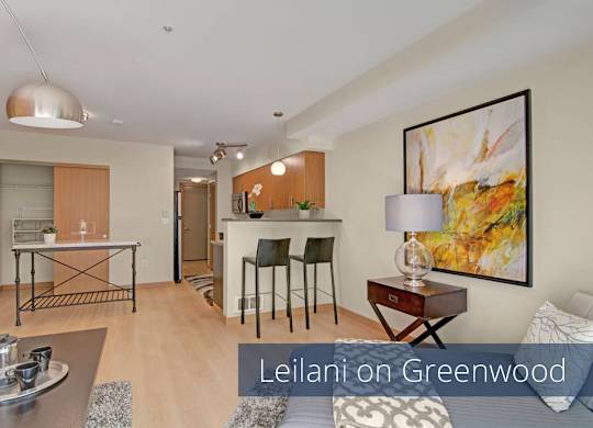 A modern open living area here at Leilani on Greenwood featuring light wood-style flooring, a cozy seating area, and contemporary artwork on neutral walls. A breakfast bar with barstool seating connects to the kitchen, while an open closet area and entry hallway add functionality to the thoughtfully designed layout.