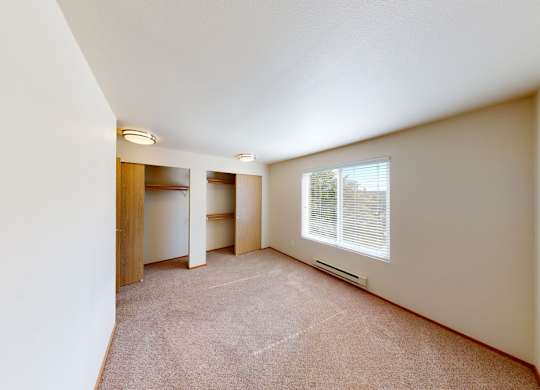 A spacious bedroom here at Leilani on Greenwood featuring soft carpeted flooring, neutral walls, and a large window with blinds that brings in natural light. Two open closets with hanging rods provide ample storage, while overhead lighting and a simple layout create a bright, comfortable sleeping space.