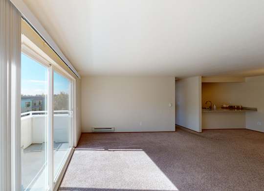 A sunlit open living area here at Leilani on Greenwood featuring soft carpeted flooring, neutral walls, and large sliding glass doors with vertical blinds that open to a private balcony. Natural light fills the space, which flows toward the kitchen area with a breakfast bar, creating an airy and inviting layout.