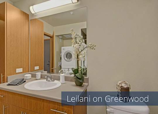 A modern bathroom here at Leilani on Greenwood featuring a spacious vanity with warm wood cabinetry, a smooth countertop with an integrated sink, and a large wall mirror beneath bright overhead lighting. The layout includes a toilet and a visible in-home laundry area reflected in the mirror, creating a functional and efficient space.
