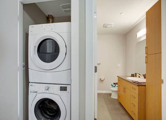 A convenient laundry nook here at Leilani on Greenwood featuring a stacked washer and dryer tucked inside a dedicated closet, positioned just off a bathroom with a long vanity, warm wood cabinetry, a smooth countertop with an integrated sink, and overhead lighting, creating a functional and efficient in-home layout.