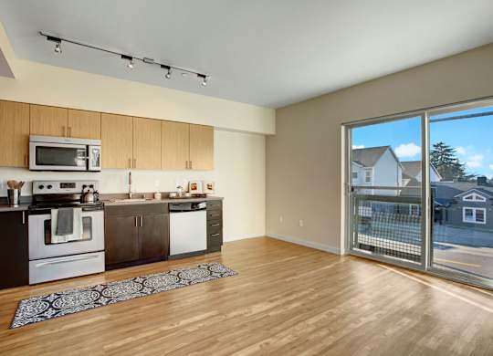 A sunlit open-concept living area here at Leilani on Greenwood featuring wood-style flooring, a modern kitchen with warm wood cabinetry, a full appliance suite, and track lighting, with large sliding glass doors that open to a private balcony and allow natural light to fill the spacious layout.