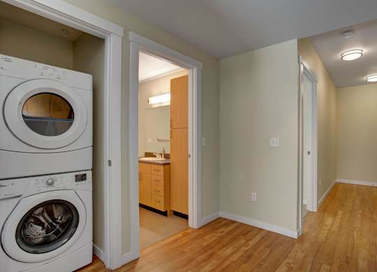 A bright interior hallway here at Leilani on Greenwood featuring wood-style flooring, neutral walls, and ceiling lighting, with an in-home stacked washer and dryer tucked into a dedicated closet and an adjacent bathroom entry showcasing warm wood cabinetry and a clean, functional layout.