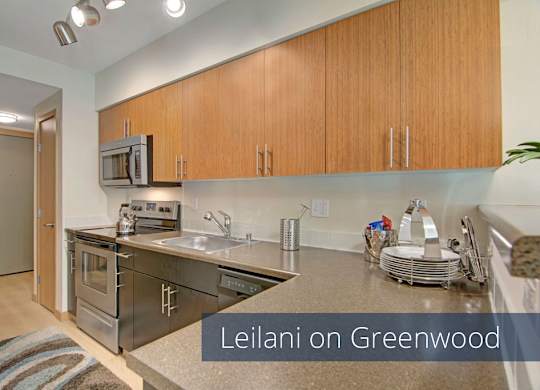 A sleek kitchen here at Leilani on Greenwood featuring flat-panel wood cabinetry, smooth countertops, and a stainless steel sink set beneath under-cabinet lighting. Dark appliances line the workspace, while light wood-style flooring and modern fixtures create a clean, contemporary cooking area.