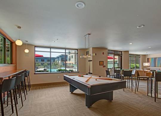 A spacious resident lounge here at Leilani on Greenwood featuring a central billiards table beneath modern pendant lighting, with surrounding bar-height seating, dining tables, and comfortable lounge furniture, all set within an open layout accented by large windows that bring in natural light and views of the outdoor courtyard.