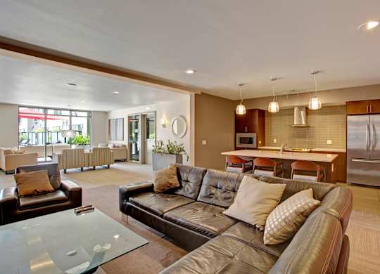 A contemporary resident lounge here at Leilani on Greenwood featuring plush seating arranged around a glass coffee table, warm neutral carpeting, and a fully equipped community kitchen with an island, bar seating, pendant lighting, and modern cabinetry, creating an inviting space for gatherings, events, and everyday relaxation.