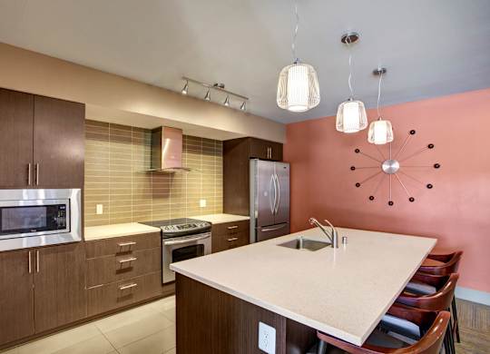 A modern community kitchen here at Leilani on Greenwood featuring an oversized island with bar seating, sleek countertops, contemporary pendant lighting, and a full appliance suite framed by dark wood cabinetry, with a tiled backsplash and bold accent wall that create a stylish space for resident gatherings and events.