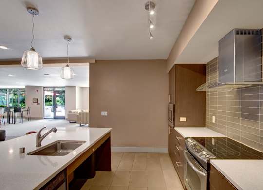 A sleek community kitchen here at Leilani on Greenwood featuring an oversized island with an integrated sink, smooth countertops, modern pendant and track lighting, and a full appliance suite with contemporary cabinetry, opening into a connected resident lounge area that creates a functional and inviting space for gatherings and events.