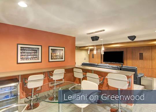 A contemporary media lounge here at Leilani on Greenwood featuring bar-height counter seating with modern stools, a glass-top table, warm accent walls with framed artwork, and a spacious viewing area with comfortable seating and a large mounted television designed for entertaining, gatherings, and movie nights.
