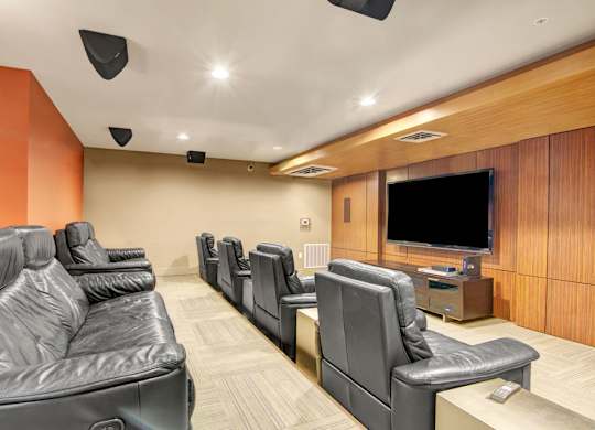 A cozy private theater room here at Leilani on Greenwood featuring rows of plush reclining leather seats, a large wall-mounted television with built-in media console, warm wood-paneled accent walls, recessed lighting, and surround sound speakers creating a comfortable space for movie nights and group viewing.