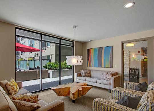 A welcoming resident lounge here at Leilani on Greenwood featuring comfortable upholstered sofas and accent chairs arranged around a sculptural wood coffee table, large floor-to-ceiling windows and sliding glass doors opening to an outdoor courtyard, modern pendant lighting, neutral carpeting, and colorful abstract wall art creating a relaxed social gathering space.