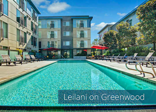 A resort-style outdoor swimming pool here at Leilani on Greenwood featuring a long rectangular pool with clear blue water, surrounding concrete pool deck, cushioned lounge chairs arranged on both sides, red patio umbrellas providing shade, landscaped planters, and modern apartment buildings enclosing the courtyard under a bright blue sky.