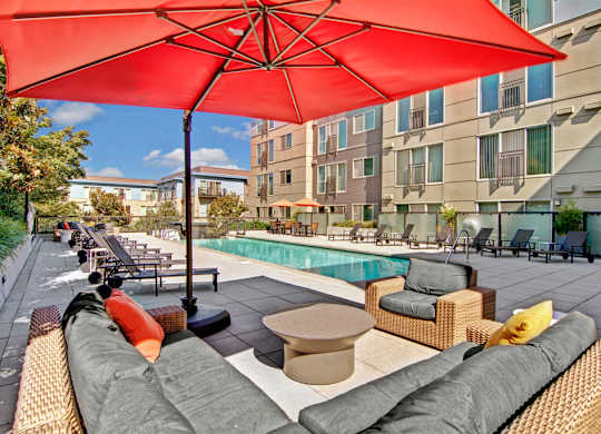 A resort-style pool courtyard here at Leilani on Greenwood featuring a long rectangular swimming pool, cushioned outdoor lounge seating in woven wicker frames, a large red patio umbrella providing shade, multiple chaise loungers lining the pool deck, modern apartment buildings surrounding the space, and landscaped greenery under a clear blue sky.