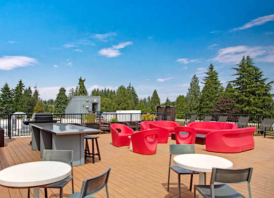 A spacious rooftop deck here at Leilani on Greenwood featuring a wood-plank patio with bright red modern lounge seating, round café tables with chairs, an outdoor grilling station, and black metal railing, all overlooking treetops and surrounding rooftops beneath a clear blue sky.